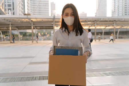Portrait of young Asian woman wearing protective face mask holding box of items after being laid off from job due toの写真素材