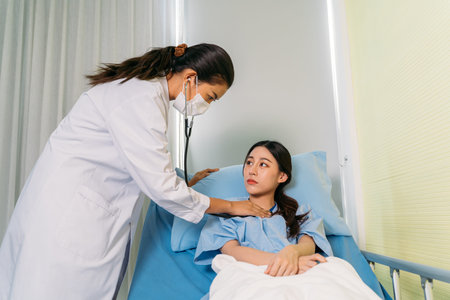 Young doctor in labcoat with face mask checking heartbeat rhythm with stethoscope of sick Asian...