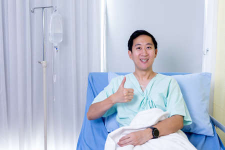 Portrait of a smiling Asian young male patient sitting on hospital bed room showing thumbs up gesture and sign to cameraの写真素材