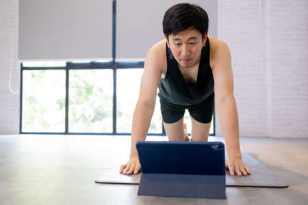 Young Asian man exercising at home while watching a video tutorial on tablet. Male fitness guy training indoors onlineの写真素材