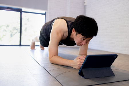 Young Asian man exercising at home while watching a video tutorial on tablet. Male fitness guy training indoors onlineの写真素材