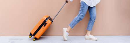 Young Asian girl happy while towing orange suitcase bumping in the air on concrete floor and desert sand colored wall background - Panorama bannerの写真素材