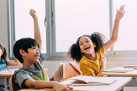 Elementary age Asian student boy raised hands up in class. Diverse group of pre-school pupils in elementary age in education building school. Volunteering and participating classroom concept.の写真素材