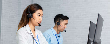 Cheerful Asian man and woman in headsets smiling and typing on computer keyboard while working in office of call centerの写真素材