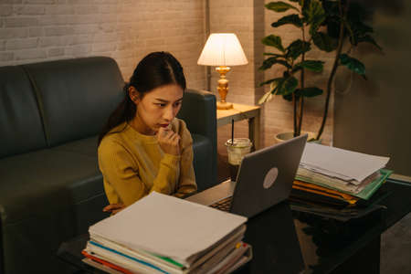 Tired young Asian female businesswoman thinking with hands on chin while staring at laptop screen with office working files on table during late night at homeの写真素材