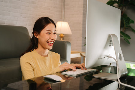 Successful happy young Asian businesswoman working from home and typing on keyboard using desktop...
