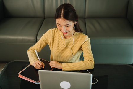 High-angle view of young Asian businesswoman attending video call with earphones and writing meeting...