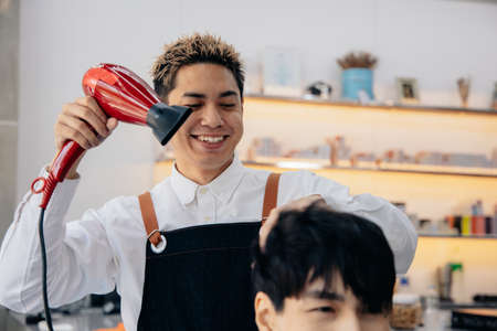 Portrait of male Asian hairdresser wearing apron working with concentration while drying handsome young mans hair in a modern barber shopの写真素材