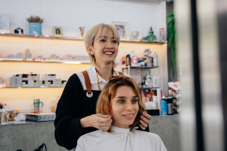 Portrait of happy young Asian woman hairstylist in apron while doing hairdressing and put care treatment while styling curly hair for clientの写真素材