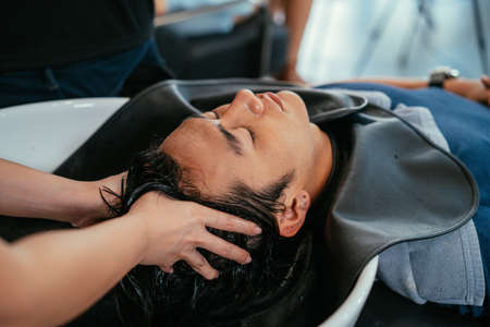 Handsome young Asian man relaxing with eyes closed with head in sink while hands of female hairdresser rinsing hair and pressing pressure pointsの写真素材