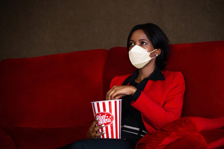 Young African American woman in formal business suit wearing a protective mask in movie theater. Shes eating a popcorn while watching - Covid-19 New Normalの写真素材