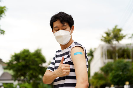 Young Asian man wearing covid-19 face protection mask showing thumbs up gesture at while looking at bandage after vaccine for protection against corona virus during dayの写真素材