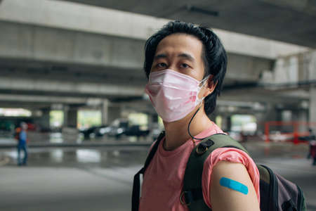 Young Asian man wearing covid-19 face protection mask looking at bandage after vaccine for protection against corona virus during day wearing a bagの写真素材