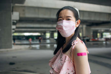 Portrait of smiling young Asian woman with covid-19 vaccination bandage standing wearing face mask for safety and precaution against coronavirus looking at cameraの写真素材