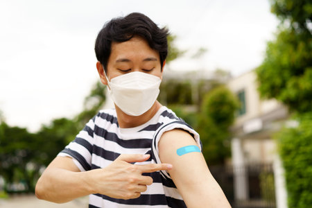 Young Asian man wearing covid-19 face protection mask pointing at bandage after receiving vaccine for protection against corona virus during dayの写真素材