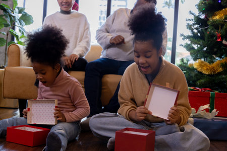 Excited two cute little african american daughters sitting on wooden floor opening christmas present...