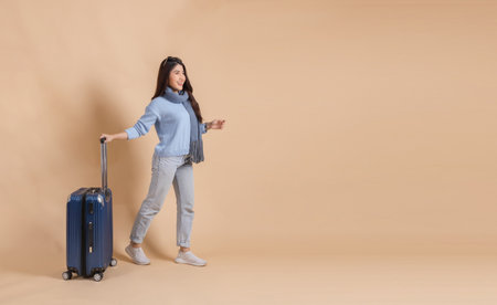 Happy Asian female tourist with luggage in blue sweater and scarf enjoying their winter vacation isolated on beige color background. Excited fashionable 20s woman going on a trip.の写真素材