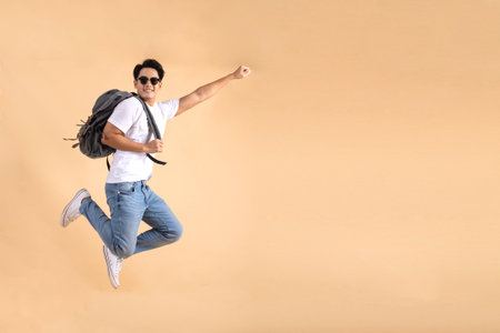 Young Asian tourist backpacker man smiling and jumping isolated on beige backgroundの写真素材