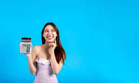 Happy smiling Asian female stylish woman holding a calculator while thinking and looking at space isolated on blue color background. Half body length studio with copy space.の写真素材