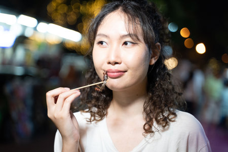 Portrait of Asian solo hipster traveller holding a fried roasted insect stick on street in Bangkok, Thailand. Female tourist exploring Southeast Asia. - copy spaceの写真素材