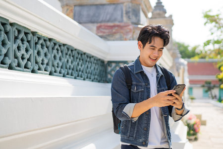Portrait of Asian male traveler using a mobile phone on sidewalk of buddhist temple on street in...
