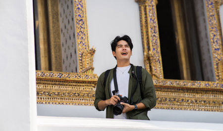 Portrait of Asian male travel photographer holding a camera in front of buddhist temple on street in Bangkok, Thailand, Southeast Asia. Handsome man taking a picture while traveling.の写真素材