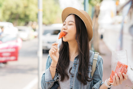 Portrait of Asian female traveler eating a slice of water melon while walking on sidewalk of buddhist temple on street in Bangkok, Thailand, Southeast Asia - Woman enjoying street food lifestyleの写真素材