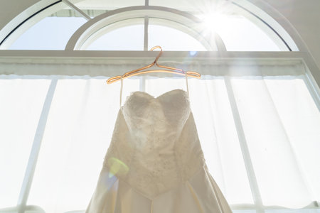 Elegant wedding dress hanging against morning light, hanging by a window in preparation of the ceremony. Symbolizing the start of a romantic wedding dayの写真素材