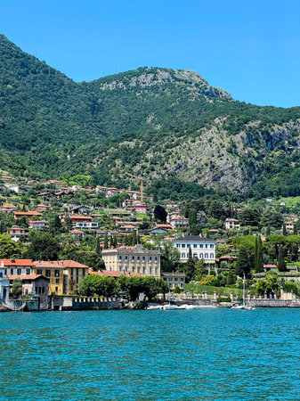 A wide view of a charming Italian town on the shores of Lake Como, with traditional buildings and villas leading down to the clear blue water, all set against a backdrop of verdantの写真素材