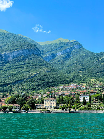 A picturesque view of the town of Bellagio on Lake Como, Italy, with its colorful buildings nestled at the foot of lush green mountains under a bright blue sky with scattered cloudの写真素材