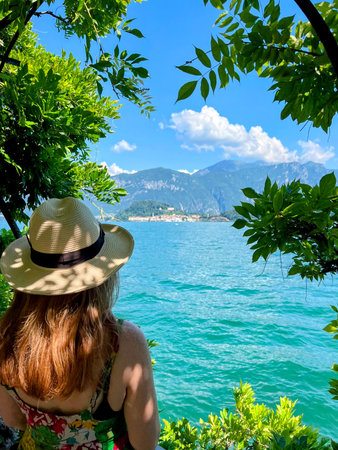 A woman in a sun hat looks out over the turquoise waters of Lake Como towards the town of Bellagio and the surrounding mountains, framed by vibrant green leaves.の写真素材