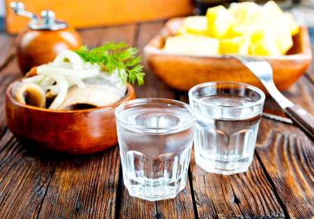 one empty second vodka-filled glass surrounded by a snack with herring and onions, a plate with fish and herbs on a wooden backgroundの写真素材