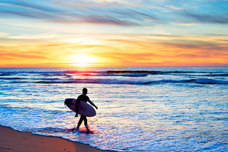 Surfer with surfboard walking on the beach at sunset