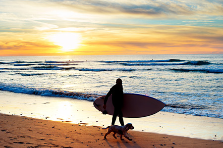 Silhouette of a surfer with a dog walking at sunset on the beachの写真素材