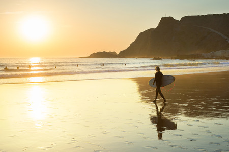 Silhouette of a woman with surfboard walking on the beach at sunset. Algarve, Portugal