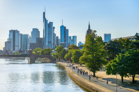 People walking by green Frankfurt embankment in evening sunlight, city skyline at background, Germanyの写真素材