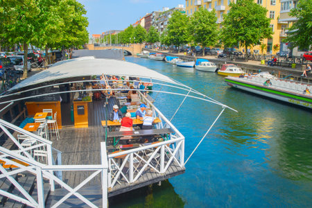 COPENHAGEN, DENMARK - JUNE 14, 2018: Sunny summer Copenhagen cityscape with green trees, tourists sitting in restaurant by canal with boatsのeditorial素材