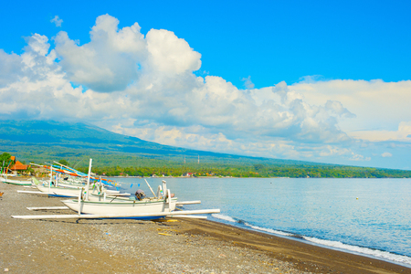 Sunshine weather at beach with traditional fishing boats, Agung volcano in clouds in background, Bali, Indonesiaの写真素材