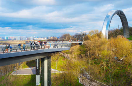 KIEV, UKRAINE - APRIL 18, 2021: People walking by new Pedestrian-Bicycle Bridge, 
People's Friendship Arch in the backgroundのeditorial素材
