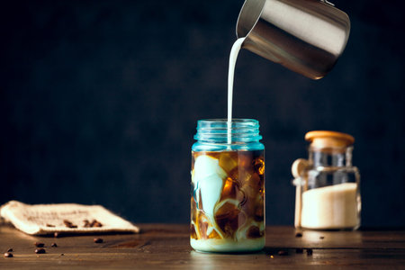 Cream Pouring Into Iced Organic Coffee Served In Blue Mason Jar On Table With Natural Sugar. Copy...