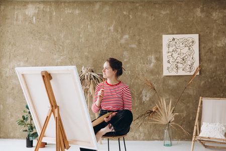 Beautiful female artist sitting near easel and looking asideの写真素材