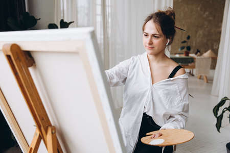 Young creative woman in earphones painting on easelの写真素材