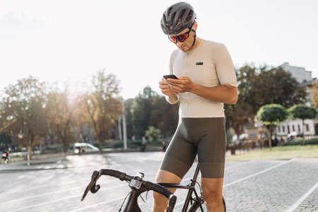 Caucasian cyclist in sportswear looking on mobile screen while standing on city street with black bike. Man taking break during workout outdoors.の写真素材