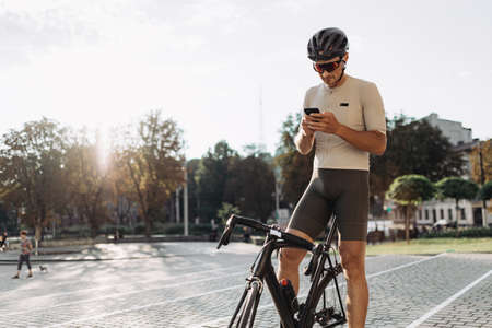 Caucasian cyclist in sportswear looking on mobile screen while standing on city street with black bike. Man taking break during workout outdoors.の写真素材