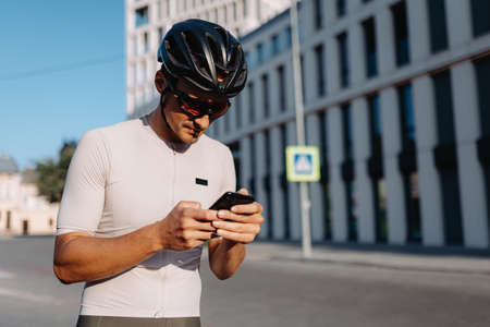 Caucasian cyclist in sportswear looking on mobile screen while standing on city street with black bike. Man taking break during workout outdoors.の写真素材