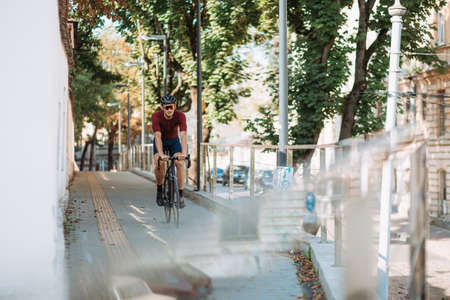 Muscular caucasian man in safety helmet and mirrored glasses using black bike for outdoors training. Cyclist spending morning time for intense workout on city street.の写真素材