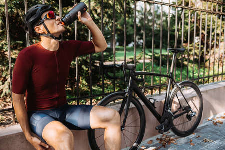 Muscular caucasian man in safety helmet and mirrored glasses using black bike for outdoors training. Cyclist spending morning time for intense workout on city street.の写真素材