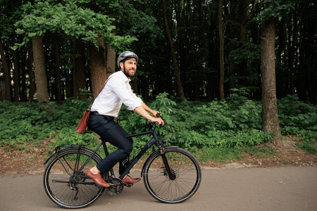 Smiling dark haired businessman on bike, riding along wooded park outdoors. Side view of man wearing...