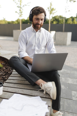 Positive office worker in headphones looking on laptop screen with joyful facial expression. Stylish male working on computer while sitting on wooden bench with leg.の写真素材