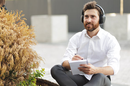 Stylish freelancer in white shirt sitting near withered thuja in urban area while noting thoughts in diary. Inspired man listening music in headphones and looking aside.の写真素材